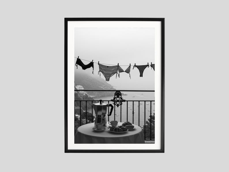Op de afbeelding: Zwart-witfoto van een balkonsc&egrave;ne. Ondergoed hangt aan een waslijn met uitzicht op een kustlandschap. Een tafel is gedekt met koffie, gebak en een koffiezetapparaat. De afbeelding is ingelijst.
