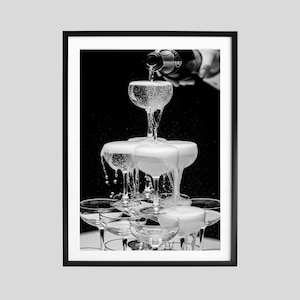 May include: A monochrome photograph showing a champagne tower being filled from a bottle. The champagne overflows, cascading down the glasses. The image is framed in a black border, creating a striking contrast against the dark background.