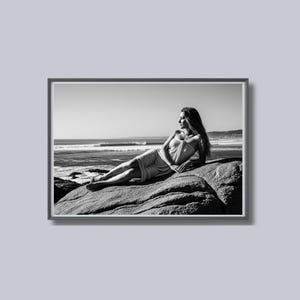 May include: Black and white framed photograph of a person reclining on a large rock at the beach. The person is wearing a light-colored garment and looking out at the ocean. The photograph is in a rectangular frame.