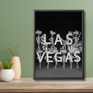 May include: Black and white framed print featuring the iconic "Las Vegas" sign with showgirls. The sign's letters are adorned with showgirls in elaborate costumes. The image evokes a vintage, retro aesthetic.
