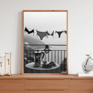 Op de afbeelding: Zwart-wit afdruk van een balkonsc&egrave;ne. Lingerie hangt aan een waslijn met uitzicht op een kustplaats en de zee. Op het balkon staat een tafel met een koffiepot, kopjes en gebak. Een decoratieve vaas en een zandloper staan op een houten oppervlak.