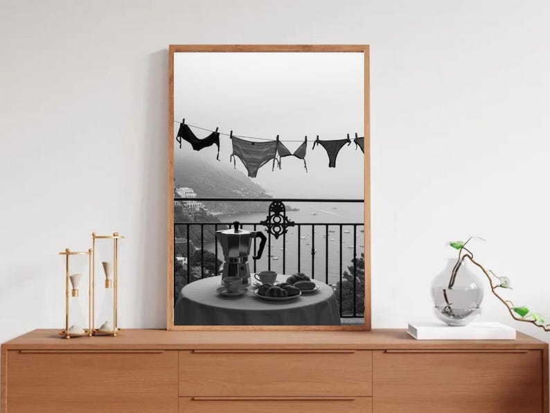 Op de afbeelding: Zwart-wit afdruk van een balkonsc&egrave;ne. Lingerie hangt aan een waslijn met uitzicht op een kustplaats en de zee. Op het balkon staat een tafel met een koffiepot, kopjes en gebak. Een decoratieve vaas en een zandloper staan op een houten oppervlak.
