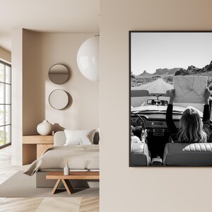 May include: Black and white framed print of a vintage convertible car on a desert road. A person in the car holds up a map. The image is displayed on a beige wall, with a bedroom interior visible on the left.