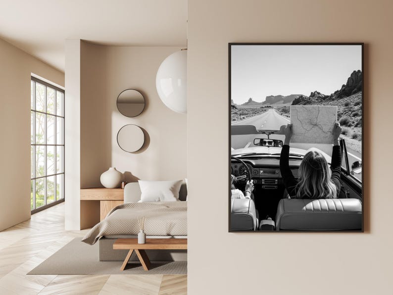 May include: Black and white framed print of a vintage convertible car on a desert road. A person in the car holds up a map. The image is displayed on a beige wall, with a bedroom interior visible on the left.