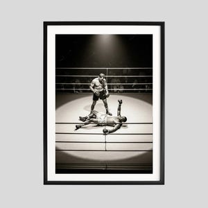 Pode incluir: Fotografia emoldurada a preto e branco de uma luta de boxe. Um pugilista está em pé sobre outro que está deitado na lona com o braço levantado. O ringue de boxe é iluminado por um holofote.