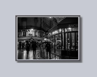 May include: Black and white photograph of a rainy street scene in Paris. People with umbrellas walk along a cobblestone street past a cafe with the name "BACIE" visible. The image captures the ambiance of a European city.