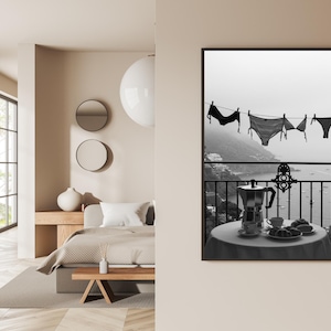 Op de afbeelding: Zwart-wit print van lingerie die aan een waslijn hangt met uitzicht op een kustlandschap. Een tafel met een koffiepot, kopjes en gebak staat op een balkon. Een serene, monochrome weergave van een ontspannen ochtend.