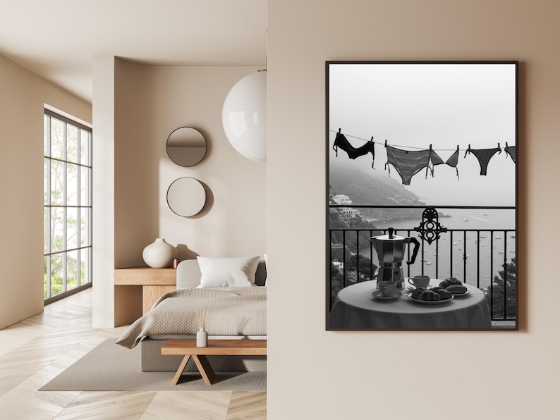 Op de afbeelding: Zwart-wit print van lingerie die aan een waslijn hangt met uitzicht op een kustlandschap. Een tafel met een koffiepot, kopjes en gebak staat op een balkon. Een serene, monochrome weergave van een ontspannen ochtend.