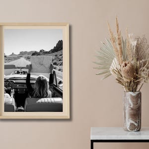 May include: Black and white framed print of a vintage convertible car on a desert road. A person in the car holds up a map. The image evokes a sense of adventure and travel. The frame is a light wood color.