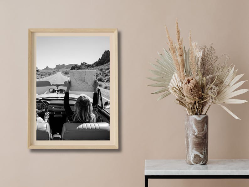 May include: Black and white framed print of a vintage convertible car on a desert road. A person in the car holds up a map. The image evokes a sense of adventure and travel. The frame is a light wood color.