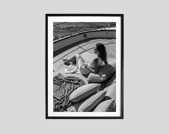 May include: Black and white framed photograph of a person reclining on a yacht deck. The person is wearing a light-colored dress and is surrounded by pillows and a blanket. The ocean is visible in the background.