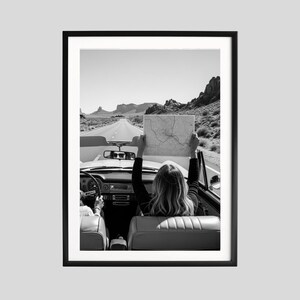 May include: Black and white framed print of a convertible car on a desert road. A person in the car holds a map up in the air. The image captures a sense of adventure and travel, with a clear sky and distant mountains.