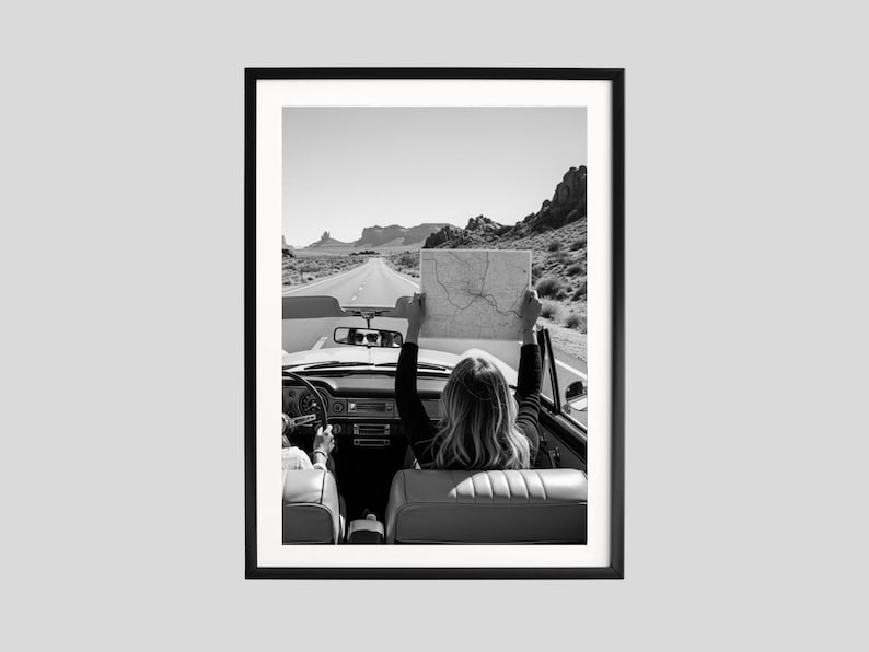 May include: Black and white framed print of a convertible car on a desert road. A person in the car holds a map up in the air. The image captures a sense of adventure and travel, with a clear sky and distant mountains.