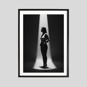 May include: Black and white framed print of a person standing in a spotlight. The person is wearing a top and pants. The image is a portrait, with the person's profile visible. The background is dark, creating a dramatic contrast with the illuminated figure.
