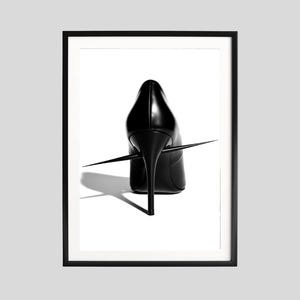 May include: Framed black and white art print featuring a black high-heeled shoe pierced by a long, sharp spike. The shoe is centered against a white background, with a shadow cast below. The artwork is enclosed in a black frame.