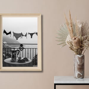 Op de afbeelding: Zwart-wit afdruk van een kustlandschap. Ondergoed hangt aan een waslijn met uitzicht op de zee. Een tafel met een koffiepot, kopjes en gebak staat op een balkon. Een vaas met gedroogde bloemen staat rechts.