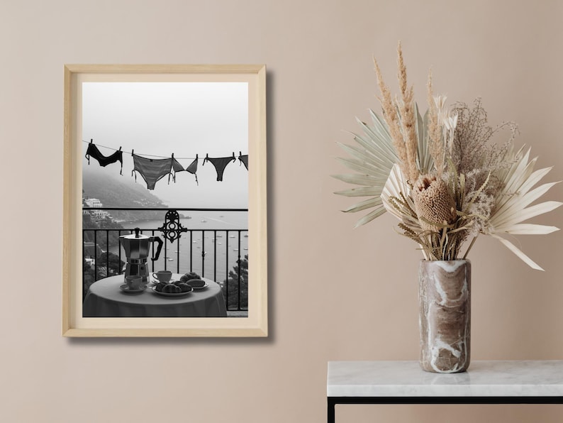 Op de afbeelding: Zwart-wit afdruk van een kustlandschap. Ondergoed hangt aan een waslijn met uitzicht op de zee. Een tafel met een koffiepot, kopjes en gebak staat op een balkon. Een vaas met gedroogde bloemen staat rechts.