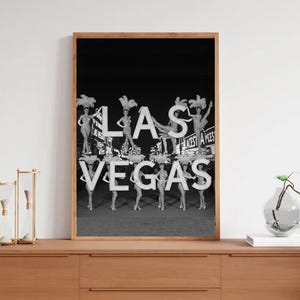 May include: Black and white framed print featuring the word "LAS VEGAS" with showgirls posing on the letters. The image has a vintage aesthetic, with the showgirls wearing elaborate headdresses and costumes. The background is a dark night sky.