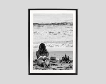 May include: Black and white framed print of a person lying on a sandy beach, facing the ocean. A hat and a small rock structure are on the sand. The ocean waves are visible in the background.