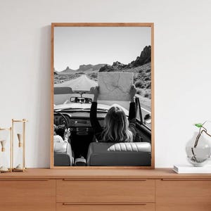 May include: Black and white framed print of a convertible car on a desert road. A person in the car holds a map up towards the sky. The image is framed in a light brown wooden frame and is displayed on a wooden dresser.