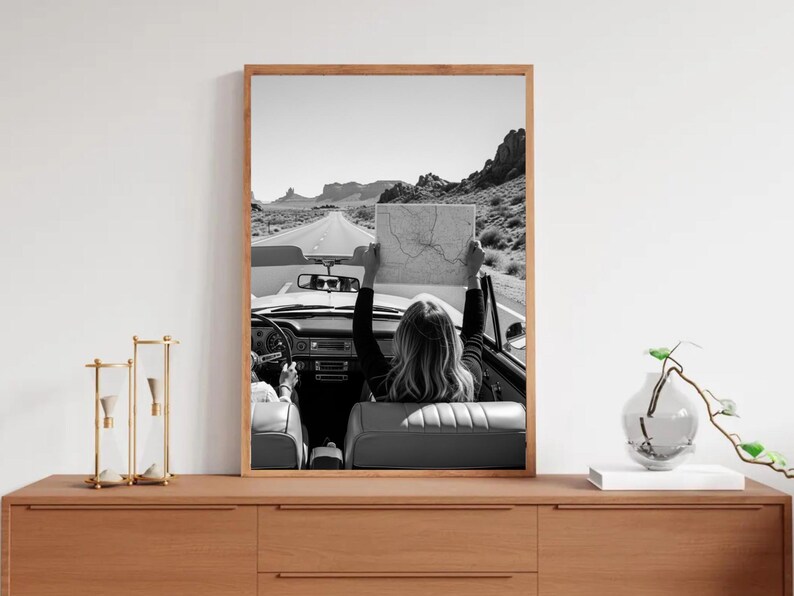 May include: Black and white framed print of a convertible car on a desert road. A person in the car holds a map up towards the sky. The image is framed in a light brown wooden frame and is displayed on a wooden dresser.