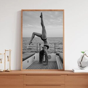 Peut inclure: Photographie en noir et blanc d'une personne faisant le poirier sur un bateau. La personne porte un bikini noir. L'arri&egrave;re-plan montre la mer et le ciel. L'image est encadr&eacute;e dans un cadre en bois.