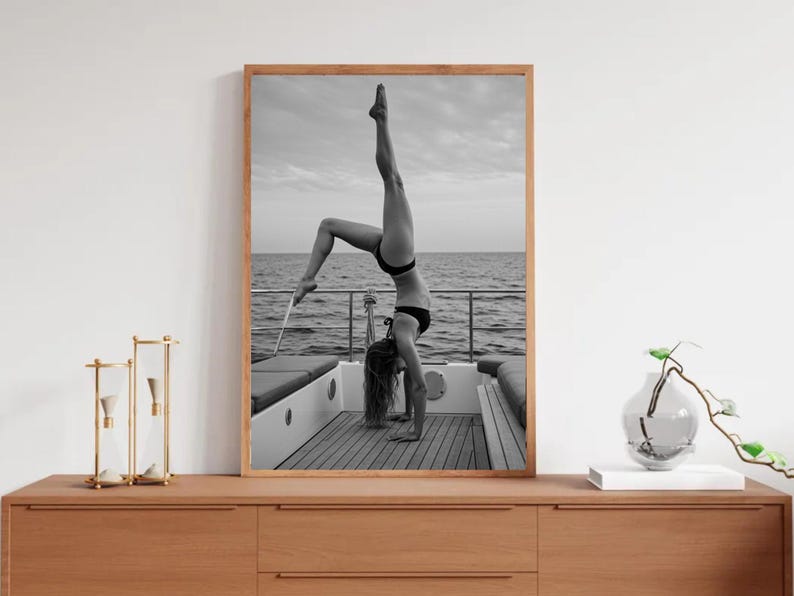 Peut inclure: Photographie en noir et blanc d'une personne faisant le poirier sur un bateau. La personne porte un bikini noir. L'arri&egrave;re-plan montre la mer et le ciel. L'image est encadr&eacute;e dans un cadre en bois.