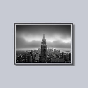 May include: Black and white framed photograph of the New York City skyline, featuring the Empire State Building as the central focus. The image captures a cloudy day, with the city's skyscrapers silhouetted against the sky.