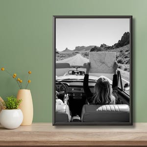 May include: Black and white framed photograph of a convertible car driving on a desert road. A person in the back seat holds up a map. The image captures a sense of adventure and travel.