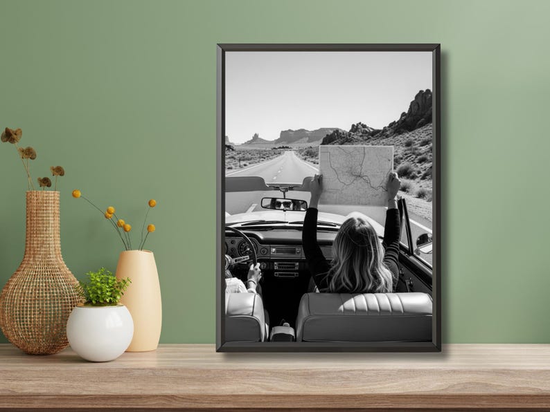 May include: Black and white framed photograph of a convertible car driving on a desert road. A person in the back seat holds up a map. The image captures a sense of adventure and travel.
