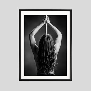 May include: Black and white framed print featuring a person with long hair, seen from the back, holding a pole with both hands above their head. The image is a close-up shot, with the person's back and arms in focus.