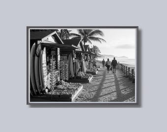May include: Black and white framed photograph of a beach scene. Several small shops line the sandy shore, displaying surfboards and various items. Palm trees and people walking along a boardwalk are visible.