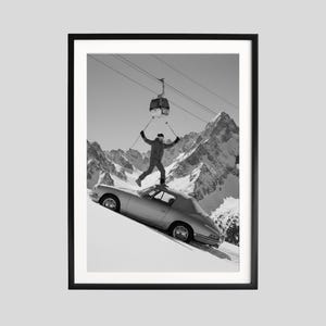 May include: Black and white framed print featuring a vintage silver car on a snowy slope. A person with skis is jumping off the car's roof, with a ski lift and snow-covered mountains in the background. The image has a retro, adventurous theme.