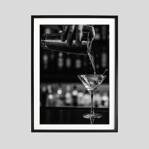 Könnte beinhalten: Schwarz-Weiß-Druck eines Martinis, der in ein Glas gegossen wird. Das Bild fängt den Moment ein, in dem ein Barkeeper einen Cocktail aus einem Shaker in ein mit einer Olive garniertes Martiniglas gießt. Der Hintergrund ist eine verschwommene Bar.