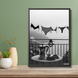 Op de afbeelding: Zwart-wit afdruk van een balkonsc&egrave;ne. Lingerie hangt aan een waslijn met uitzicht op de kust. Een tafel is gedekt met een koffiepot, kopjes en gebak. De sc&egrave;ne roept een ontspannen vakantiesfeer op.