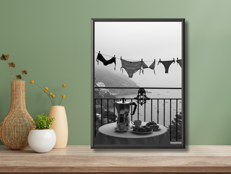 Op de afbeelding: Zwart-wit afdruk van een balkonsc&egrave;ne. Lingerie hangt aan een waslijn met uitzicht op de kust. Een tafel is gedekt met een koffiepot, kopjes en gebak. De sc&egrave;ne roept een ontspannen vakantiesfeer op.