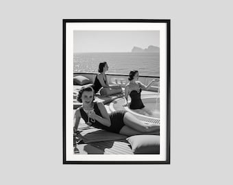 May include: Black and white framed print of three women on a yacht. One woman reclines, holding a cocktail, while others relax on deck and in a small pool. The sea and mountains are visible in the background.