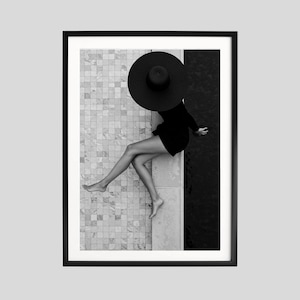 May include: Black and white framed art print. A person wearing a black hat and dress sits by a pool. The image is a high-angle shot, with the person's legs extended. The background is a tiled surface and dark water.