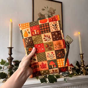 May include: A rectangular fabric book cover featuring a patchwork design of autumn-themed squares in shades of orange, green, red, and brown. The pattern includes illustrations of pumpkins, mushrooms, acorns, leaves, and plaid. It has a green ribbon tie.