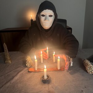 May include: A person wearing a white mask and a black hooded sweatshirt sits at a table surrounded by lit candles. A wooden incense holder and bundles of sage are also on the table, creating a mystical atmosphere.
