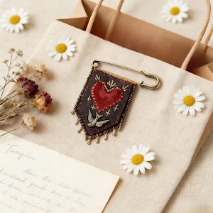 Puede incluir: Broche decorativo con un corazón rojo y un diseño de pájaro sobre un fondo negro, sujeto con un imperdible grande. El broche está adornado con detalles dorados y pequeños abalorios colgantes. La imagen incluye margaritas y una bolsa de papel.