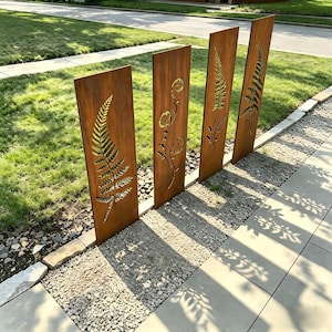 Könnte beinhalten: Vier verwitterte Gartenpaneele aus Metall mit ausgeschnittenen Farn- und Blumendesigns. Die rechteckigen Paneele stehen aufrecht in einem Kiesbett und werfen Schatten auf den Beton-Bürgersteig. Die Paneele haben eine natürliche, rostbraune Farbe.