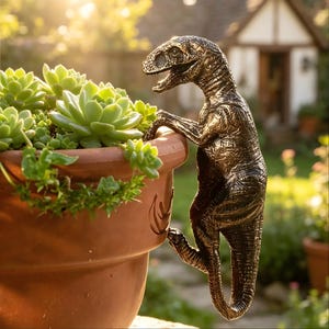 Op de afbeelding: Een bronzen dinosaurussculptuur die een terracotta bloempot beklimt, gevuld met groene vetplanten. De sculptuur is gedetailleerd en gestructureerd, met de kop en armen van de dinosaurus die over de rand van de pot reiken.