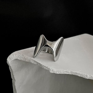Peut inclure: Une bague argentée polie au design abstrait unique. La bague présente une forme lisse et incurvée avec une indentation centrale, posée sur une surface blanche et texturée sur fond noir. Le design de la bague est moderne et minimaliste.