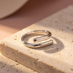 Peut inclure: Une bague argentée au design minimaliste. La bague présente une finition lisse et polie et un style enveloppant unique. La bague est présentée sur une surface texturée et de couleur claire, mettant en valeur son élégance simple. Un bijou moderne.
