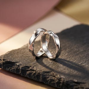 Peut inclure: Une bague argentée au design fluide et entrelacé. La bague a une surface polie et réfléchissante et repose sur une surface sombre et texturée. La forme de la bague est organique et fluide, avec des courbes douces et une esthétique moderne.