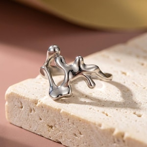 Peut inclure: Une bague argentée présentant une forme organique irrégulière rappelant du métal fondu avec des surfaces lisses et réfléchissantes. Le design se compose de formes fluides interconnectées créant une structure ouverte et abstraite.