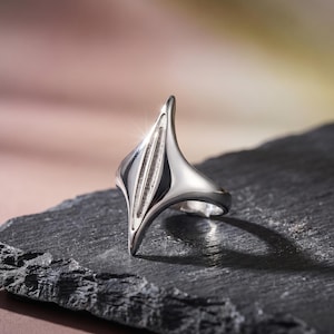 Peut inclure: Une bague en argent poli au design abstrait unique. La bague présente une forme centrale allongée avec des rainures verticales, flanquée de côtés incurvés évasés. Elle est présentée sur une surface sombre et texturée, soulignant ses qualités réfléchissantes.