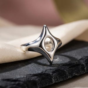 Peut inclure: Une bague en argent poli présentant une forme d'étoile à quatre branches avec une surface lisse et arrondie et une découpe ovale centrale. Le métal a une finition réfléchissante, soulignant les bords incurvés et le design ouvert de l'étoile.