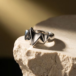 Peut inclure: Une bague en argent au design angulaire unique. La bague présente une série de formes en zigzag pointues qui créent un look moderne et accrocheur. Le métal poli reflète la lumière, soulignant les détails complexes de la bague. La bague est présentée sur une surface texturée et de couleur claire.